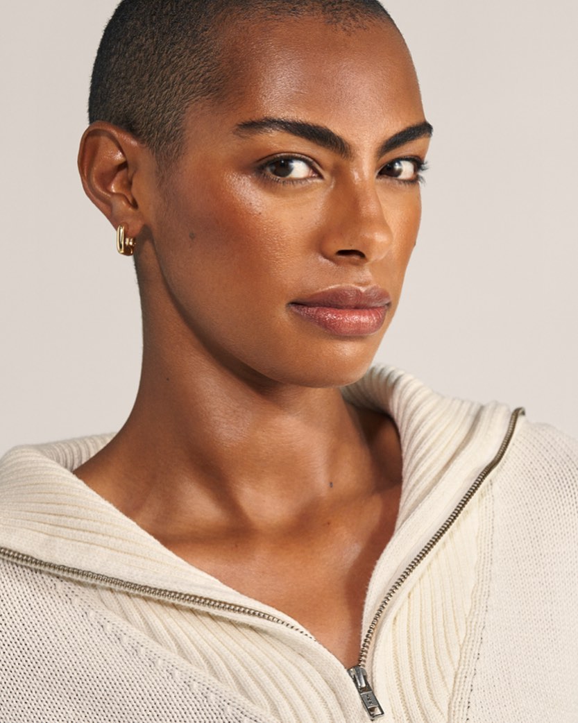 Close-up shot of an adult woman with short hair and gold earrings, wearing a white knitted sweater with a zipper in a studio setting.