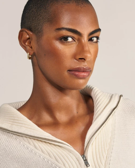 Close-up shot of an adult woman with short hair and gold earrings, wearing a white knitted sweater with a zipper in a studio setting.