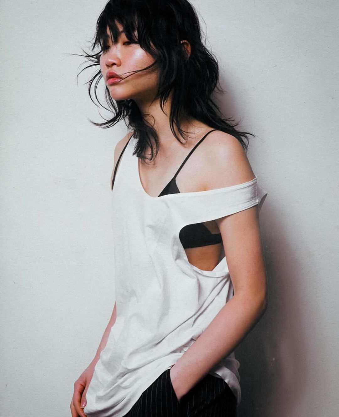 Portrait of an Asian woman with dark wavy hair wearing a white top and pinstripe pants posing indoors against a white wall.