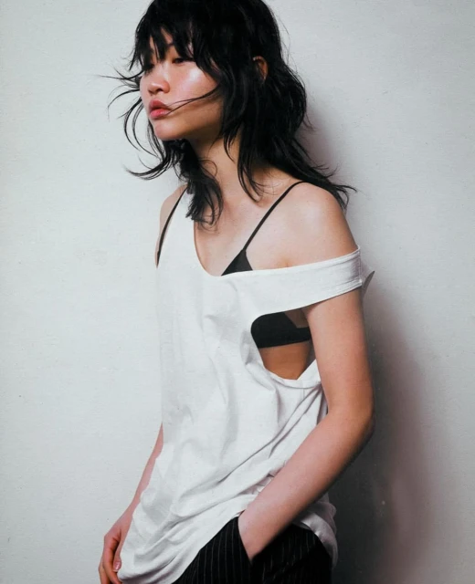Portrait of an Asian woman with dark wavy hair wearing a white top and pinstripe pants posing indoors against a white wall.