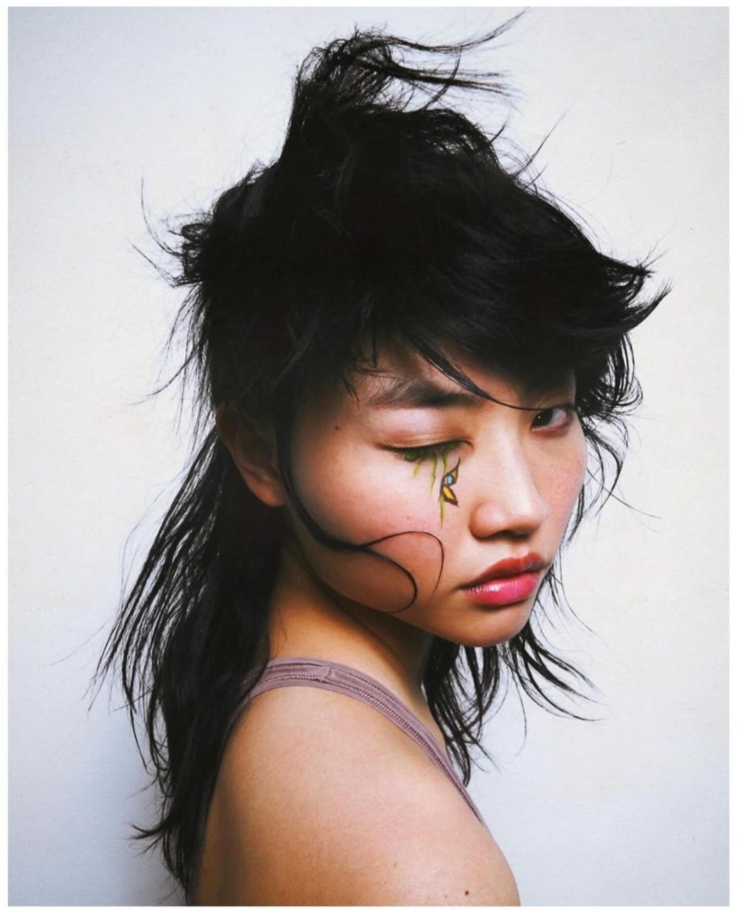 Portrait of a young Asian woman with stylized makeup, eyes closed, black hair, and light skin tone against a plain white background.