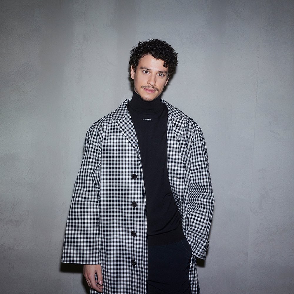 Adult male in a black turtleneck and a black and white gingham coat standing against a light grey wall in a portrait orientation.