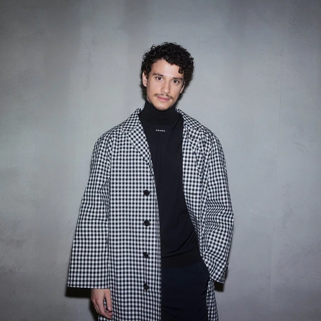 Adult male in a black turtleneck and a black and white gingham coat standing against a light grey wall in a portrait orientation.