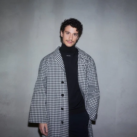 Adult male in a black turtleneck and a black and white gingham coat standing against a light grey wall in a portrait orientation.