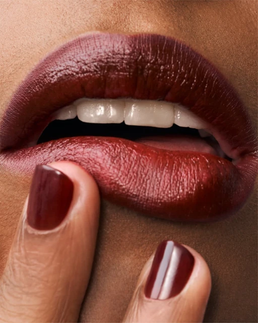 Close-up of a person with dark skin and red lipstick touching their lower lip with their finger, showcasing glossy nails.