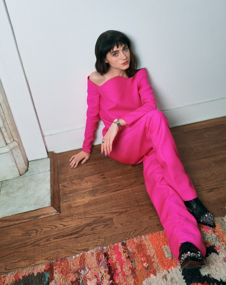 A brunette woman with short hair is posing while wearing a pink long sleeve top and pants with heeled booties indoors against a white wall.
