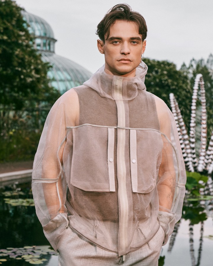 Adult male model in sheer hooded jacket, posing outdoors in garden with pool and glass conservatory in the background on a cloudy day.