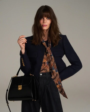 Model in navy jacket holding black leather handbag against a neutral grey studio background by Emily Soto