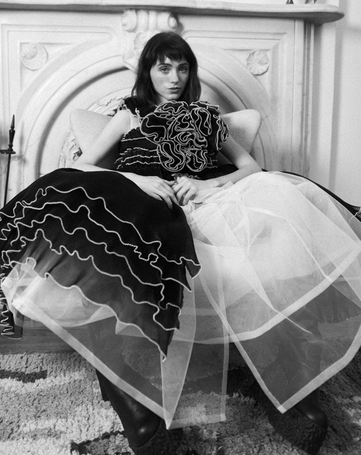 Model wearing a voluminous ruffled black and white dress seated in an armchair in front of an ornate fireplace.