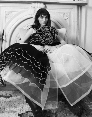 Model wearing a voluminous ruffled black and white dress seated in an armchair in front of an ornate fireplace.