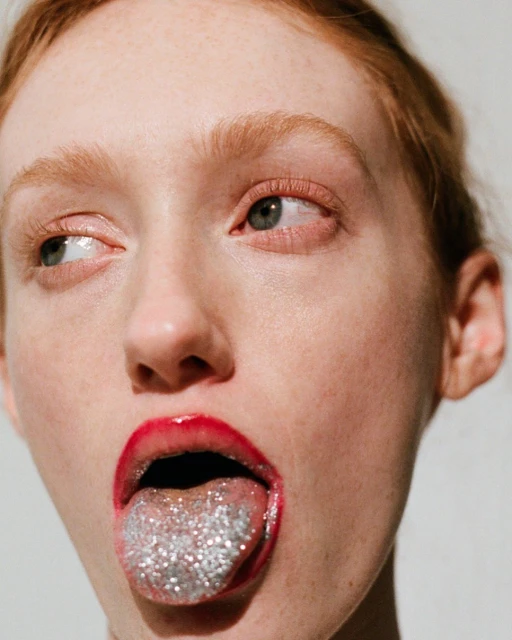 Close-up of a person with red lipstick sticking their tongue out, which is covered in silver glitter, against a white background.