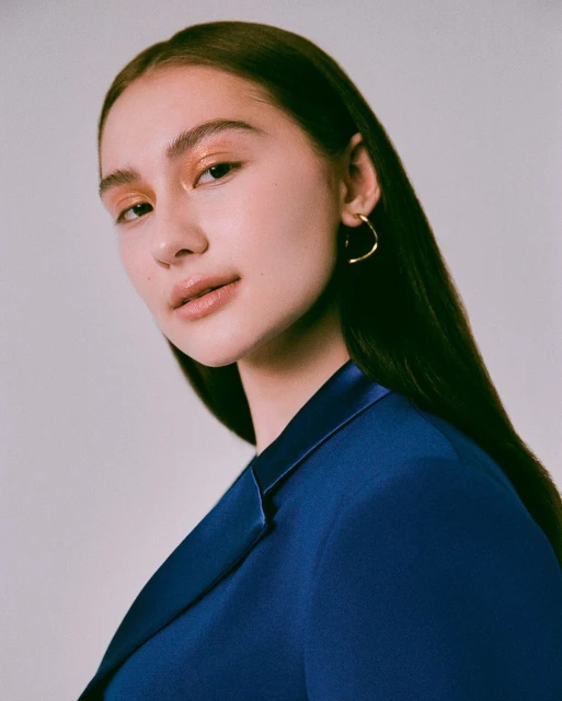 Portrait of a young woman with golden eyeshadow wearing a blue blazer and hoop earring, set against a neutral background.