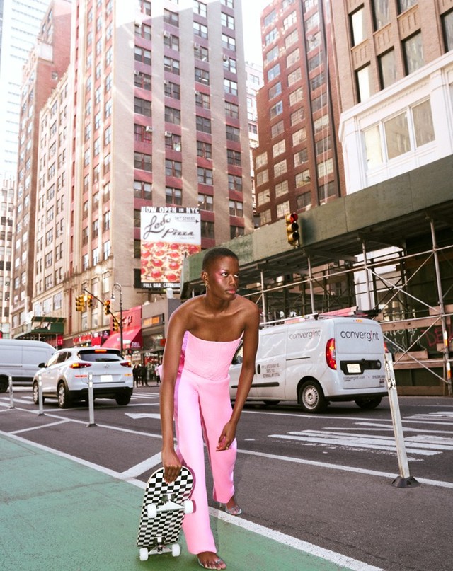 Young adult woman in a pink suit poses on a city street holding a skateboard, with buildings, cars, and vans in the background.