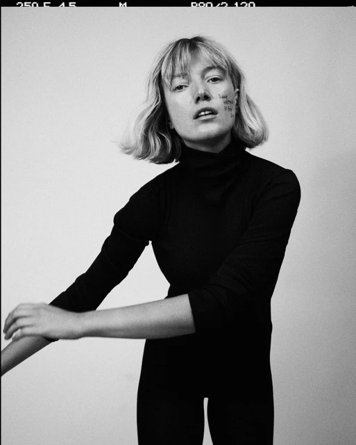 Black and white portrait of a young adult woman with bob haircut, wearing a dark turtleneck, posing in a photo studio against a grey background.