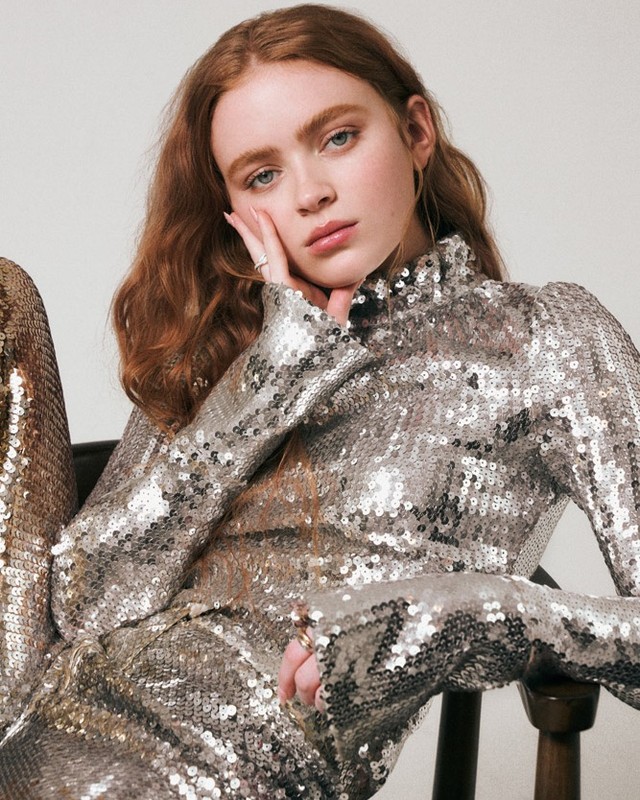 A poised young woman with red hair, wearing a shimmering silver sequined top and pants, rests her face in her hand with a neutral expression.