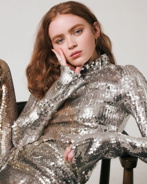 A poised young woman with red hair, wearing a shimmering silver sequined top and pants, rests her face in her hand with a neutral expression.