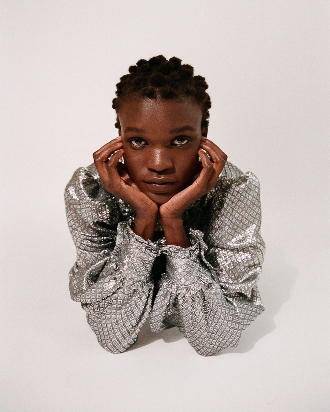 Eye-level portrait of a young Black woman in a silver patterned top with hands supporting her face against a white studio backdrop.