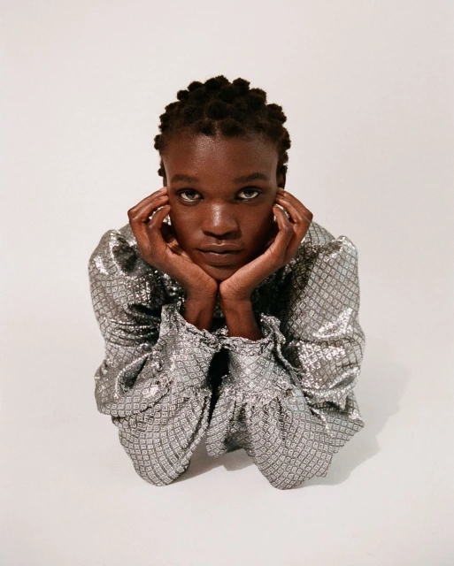 Eye-level portrait of a young Black woman in a silver patterned top with hands supporting her face against a white studio backdrop.