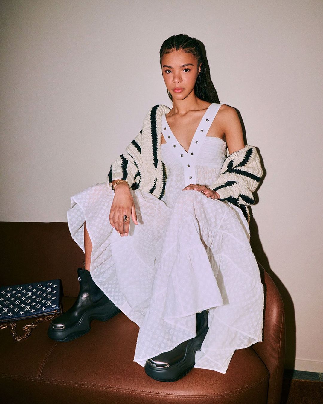 A young adult female model wearing a white dress and cardigan sits on a brown chair next to a handbag in an indoor setting.