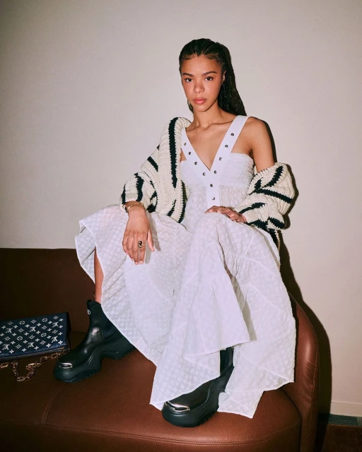 A young adult female model wearing a white dress and cardigan sits on a brown chair next to a handbag in an indoor setting.