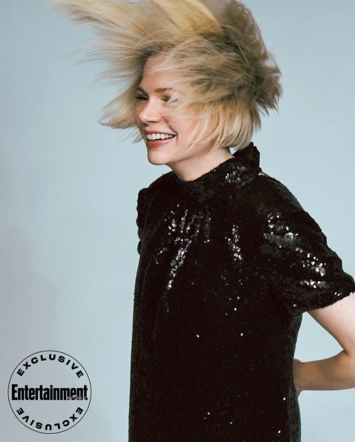 Smiling blonde woman in a black sequin dress with a light blue background. Her hair is styled upwards in a dynamic fashion look.