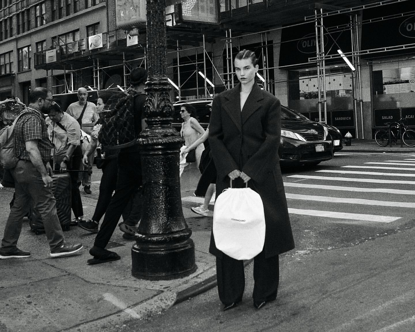 A model stands on a city street holding a white drawstring bag, wearing a dark coat and pants, with pedestrians and traffic in the background.