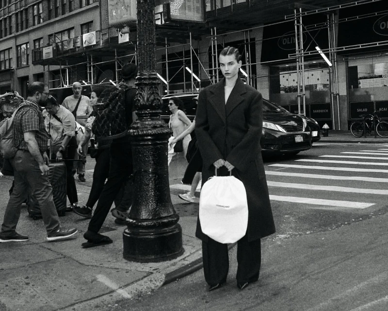 A model stands on a city street holding a white drawstring bag, wearing a dark coat and pants, with pedestrians and traffic in the background.