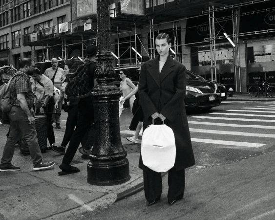 A model stands on a city street holding a white drawstring bag, wearing a dark coat and pants, with pedestrians and traffic in the background.