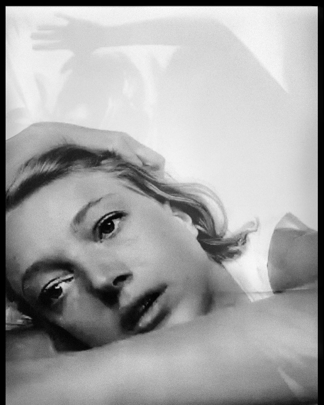 Monochrome portrait of a fair-skinned adult woman with her hand on her head, laying down, eyes open, soft light, and a shadow in background.