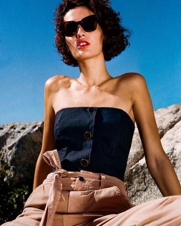 Woman in dark sunglasses wearing a strapless blue top and beige trousers against rock formations and a clear blue sky