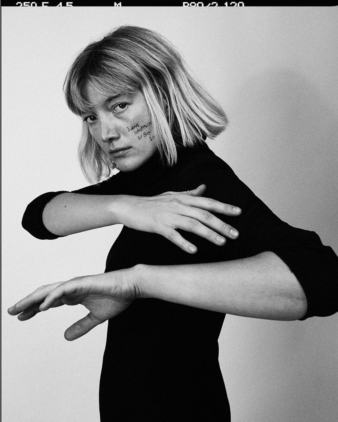 Portrait of a young adult woman with blonde hair and writing on her face, wearing a black turtleneck against a white wall.
