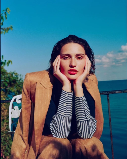 Portrait of a woman in a tan suit sitting with her hands on her face in front of a blue sky and ocean background on a sunny day.