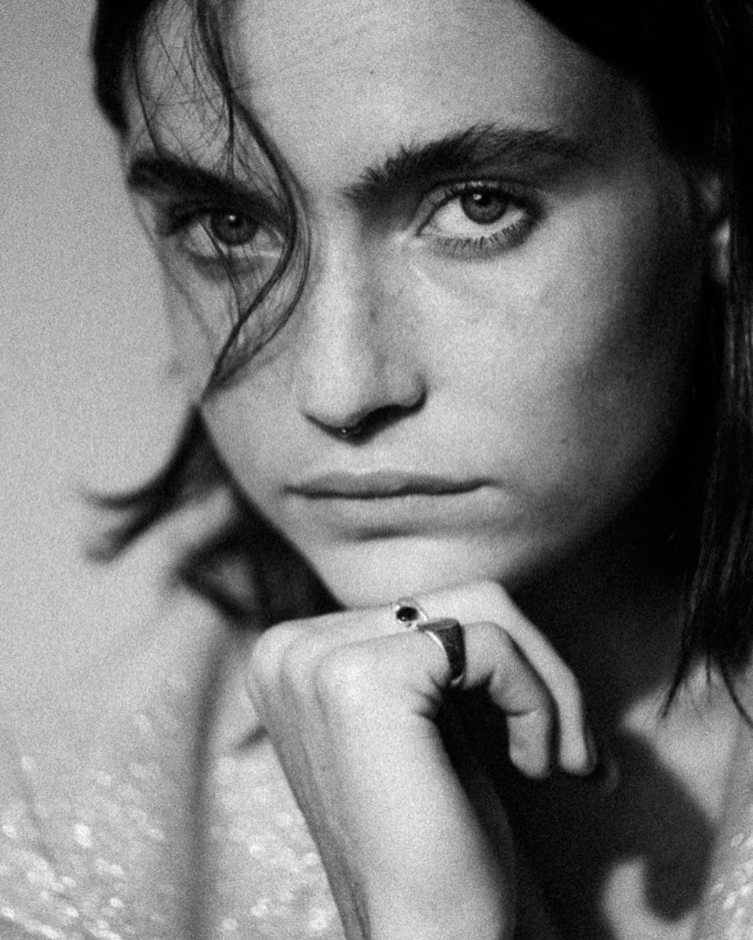 Black and white portrait of a fair-skinned young adult woman with dark hair, looking directly at the camera, hand under chin, ring visible.
