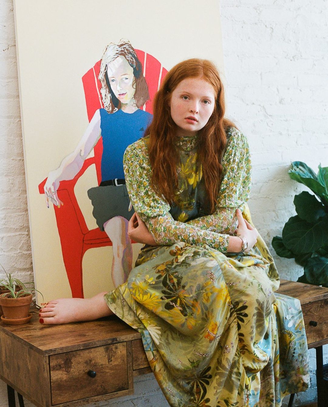 Portrait of a red-haired woman in a floral dress sitting on a wooden desk in front of a painting in a bright indoor setting.