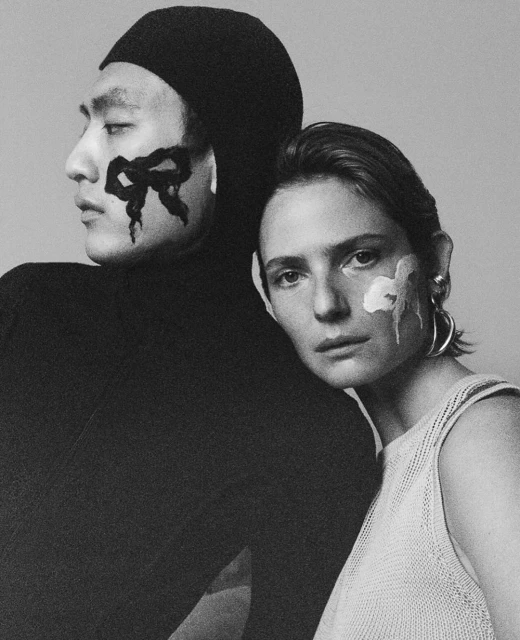 Monochrome studio portrait of two fashion models with face paint: an Asian male in a balaclava and a Caucasian female with silver hoop earrings.
