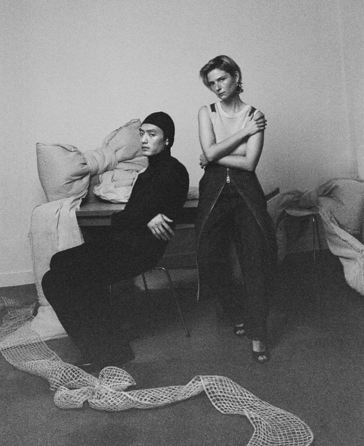 Black and white fashion shot: Asian male in black beanie sitting on a chair, Caucasian woman standing with arms crossed indoors.