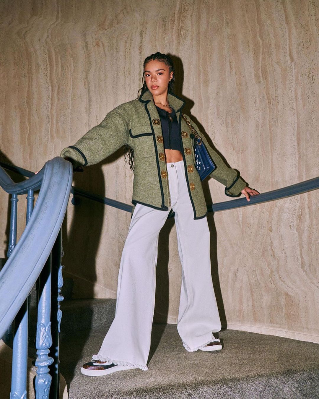 Stylish woman in a green jacket and wide-leg pants poses on a stairway with a marble wall, showcasing a blend of fashion and architecture.