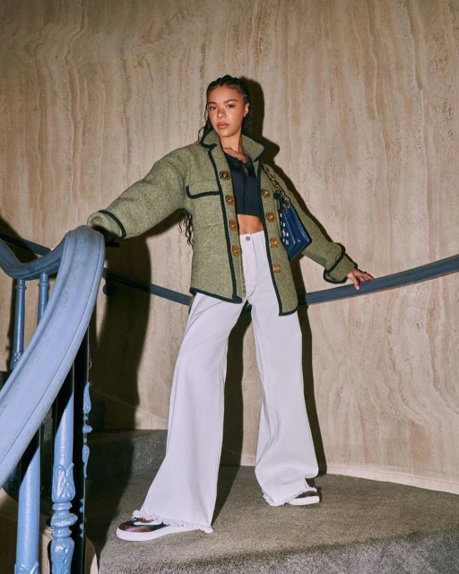 Stylish woman in a green jacket and wide-leg pants poses on a stairway with a marble wall, showcasing a blend of fashion and architecture.