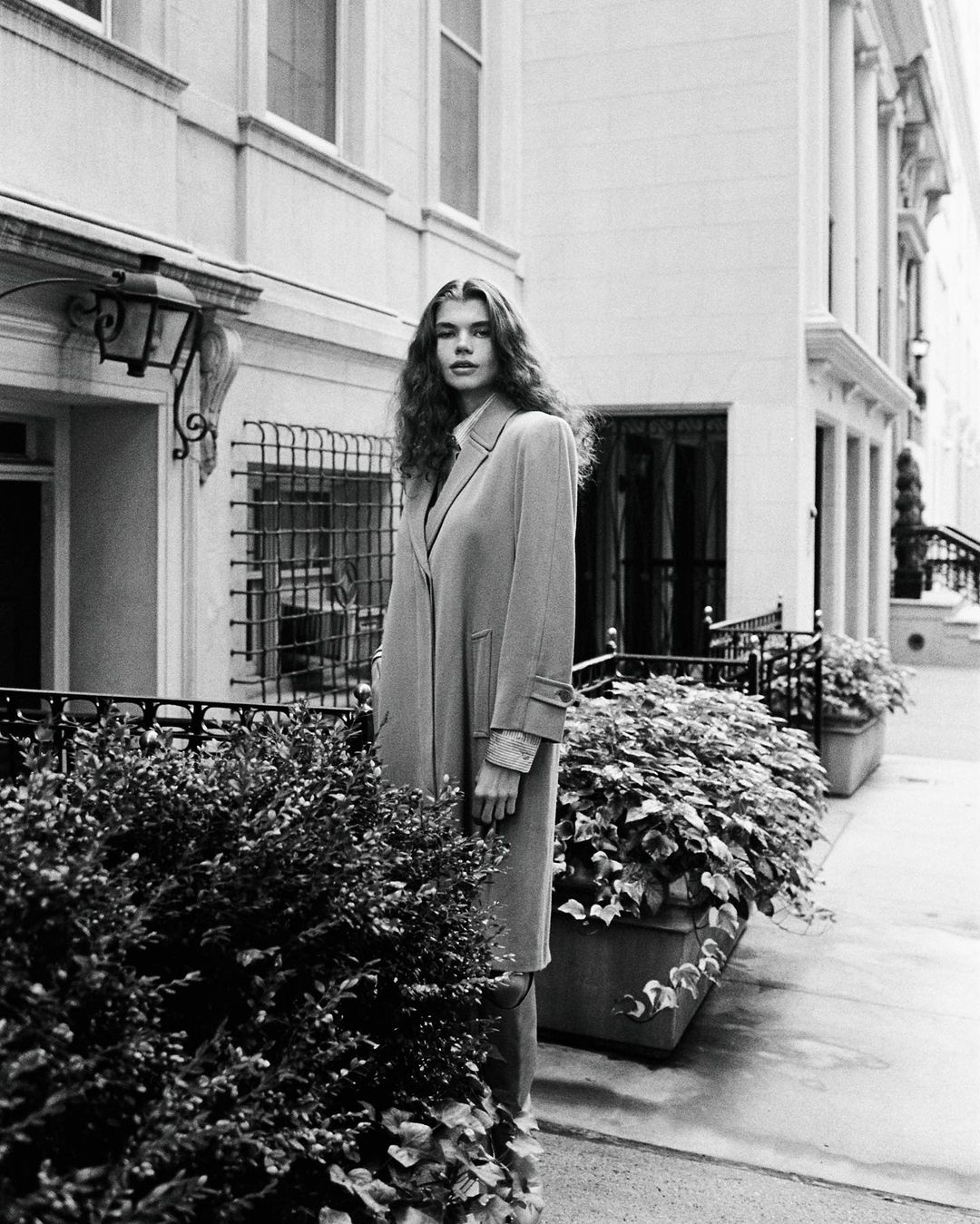 Monochrome shot of a woman with long wavy hair in a long coat standing near a hedge in front of a city building, photographed at eye-level.