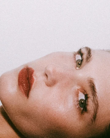 Close-up portrait of an adult woman lying down, featuring red lipstick, fair skin, and direct eye contact against a white background.