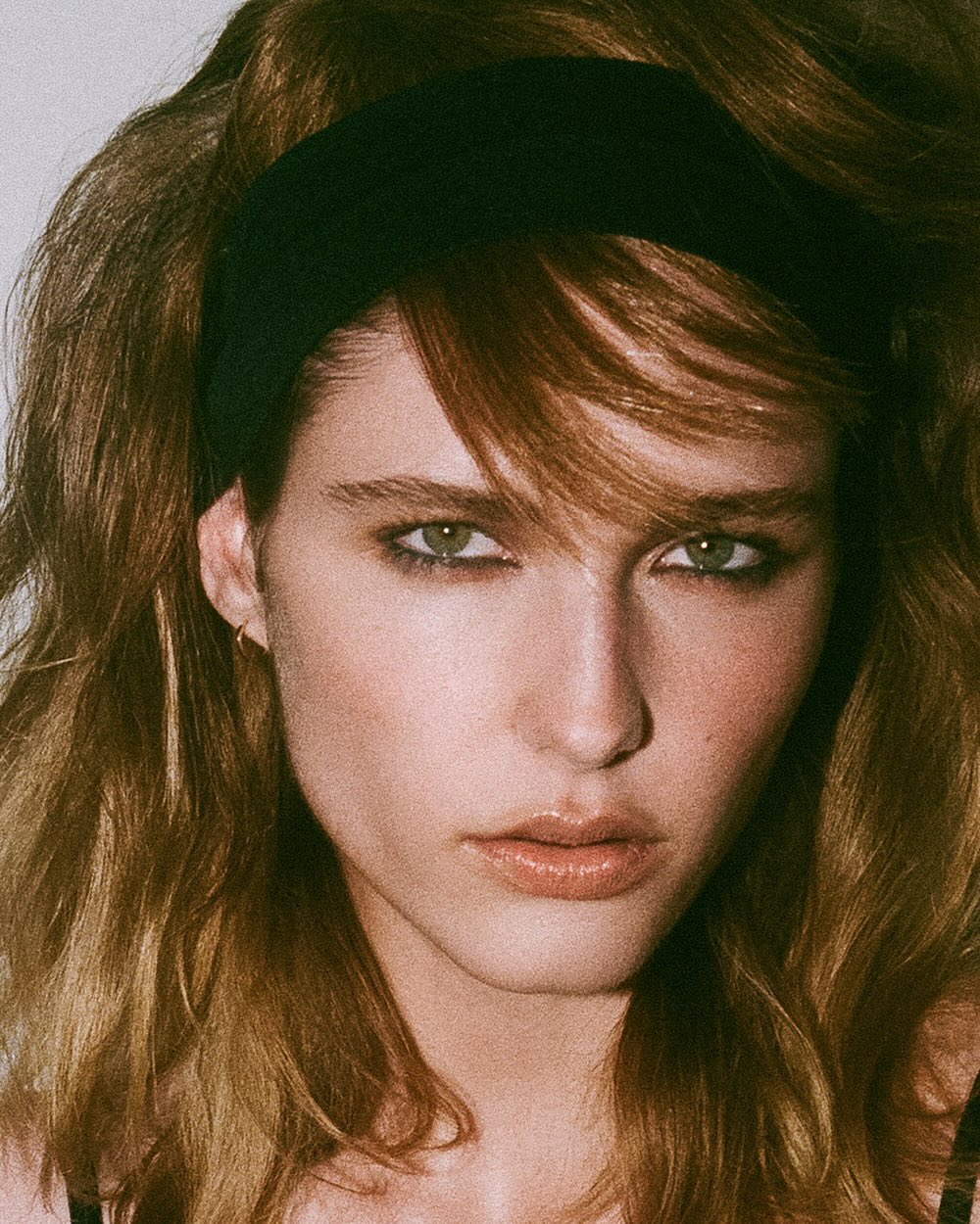 Portrait of a young adult woman with wavy brown hair, wearing a black headband, posing in a close-up shot against a plain white background.