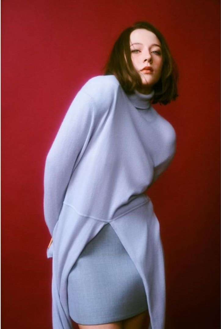 Woman in light blue turtleneck sweater and skirt poses against a red background in a fashion studio portrait with soft, diffused lighting.