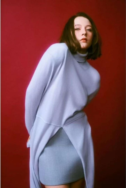 Woman in light blue turtleneck sweater and skirt poses against a red background in a fashion studio portrait with soft, diffused lighting.