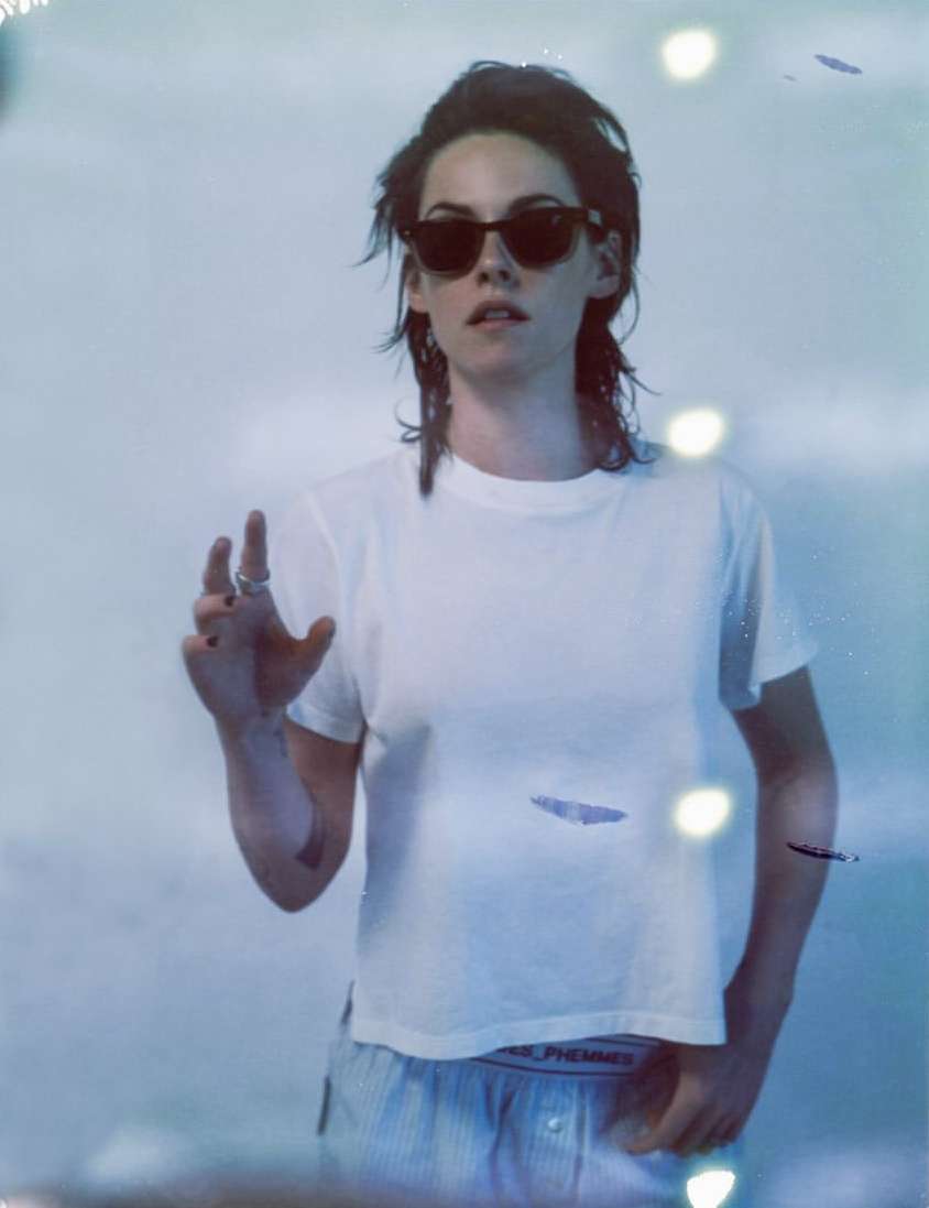 Portrait of a woman with sunglasses and a white t-shirt, standing in front of a bright white background, indoor lighting and blue hues.