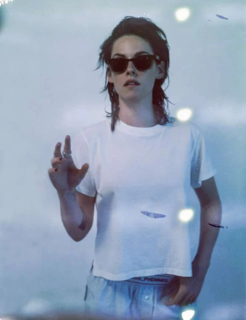Portrait of a woman with sunglasses and a white t-shirt, standing in front of a bright white background, indoor lighting and blue hues.