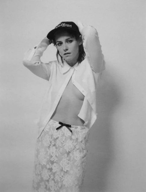 Black and white photo of a woman wearing a baseball cap and a partially unbuttoned white shirt and floral lace skirt in a studio setting.