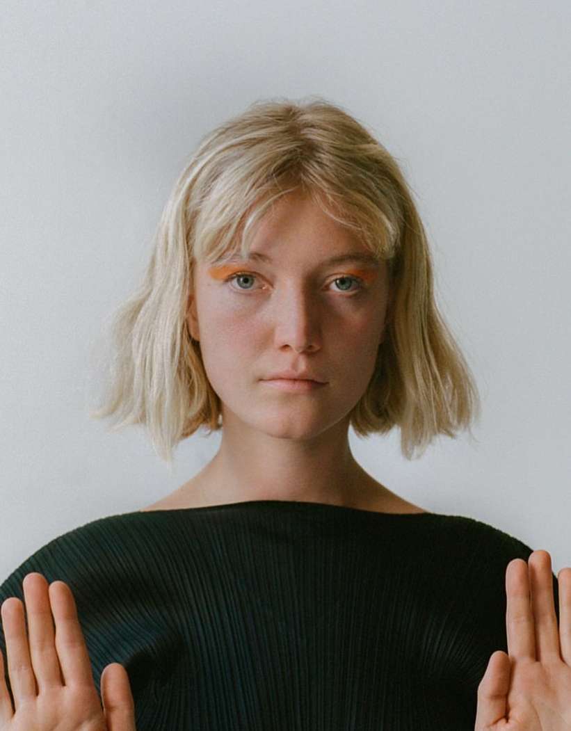 Portrait of a blonde woman with orange eyeshadow wearing a black top against a white background