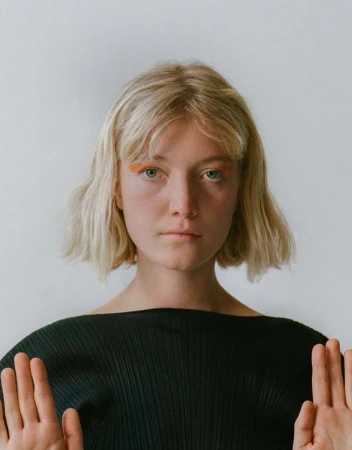 Portrait of a blonde woman with orange eyeshadow wearing a black top against a white background