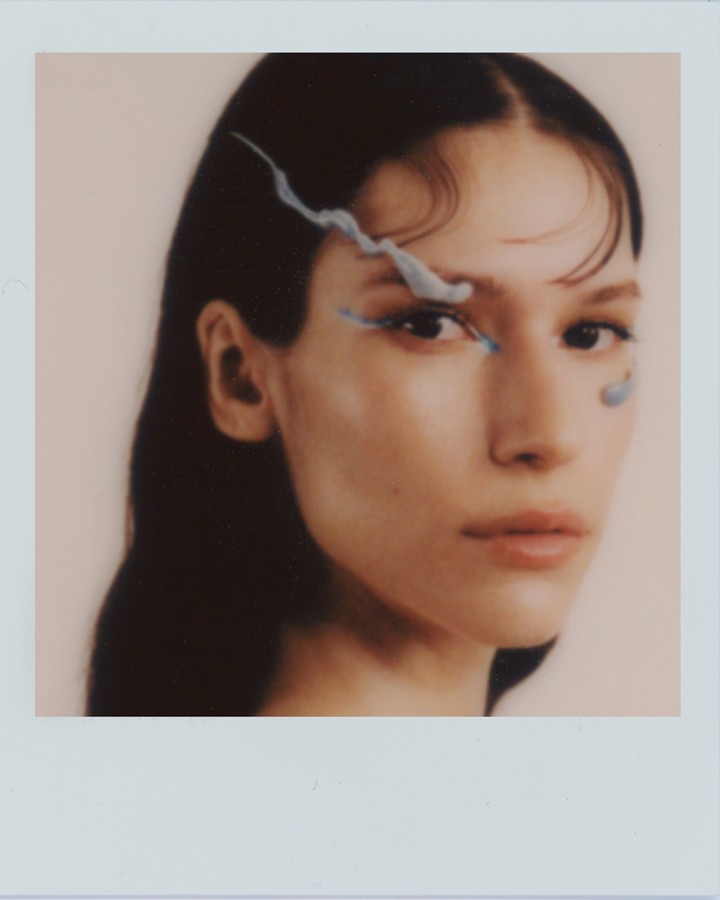 Close-up studio shot of an adult woman with dark hair and blue makeup accenting her eye, captured in soft focus and muted lighting.