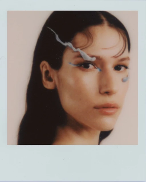 Close-up studio shot of an adult woman with dark hair and blue makeup accenting her eye, captured in soft focus and muted lighting.